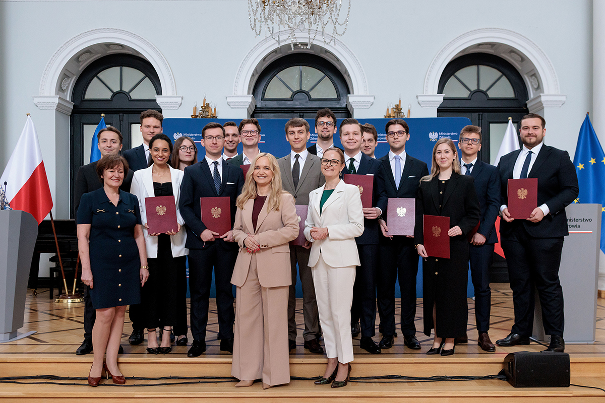 Grupa kilkunastu osób stoi w dwóch rzędach w sali reprezentacyjnej. Większość osób trzyma bordowe teczki z godłem. W centrum stoją osoby uczestniczące w oficjalnej części wydarzenia. W tle widoczne są flagi Polski i Unii Europejskiej oraz ścianka z logotypami Ministerstwa Zdrowia.