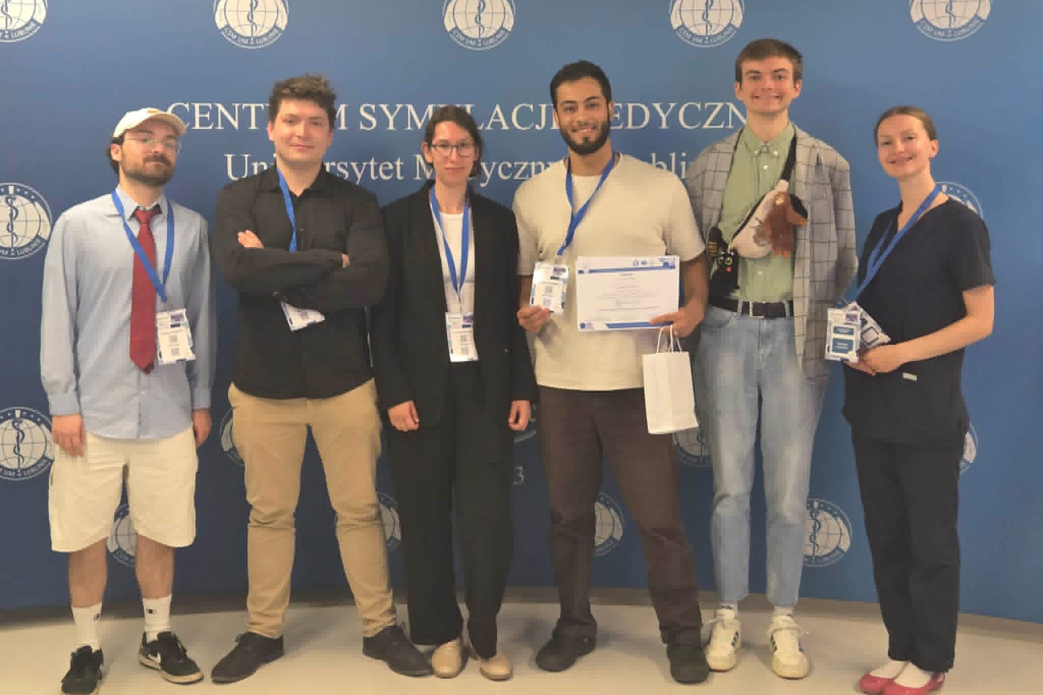 Six people are standing in a row against a blue backdrop with the inscription “Medical Simulation Center, Medical University of Lublin” and repeating logos. All of them have ID badges on blue lanyards around their necks. One person is holding a diploma and a gift bag, while another is holding a plush toy attached to a bag. The remaining people are standing next to each other, posing for a group photo.
