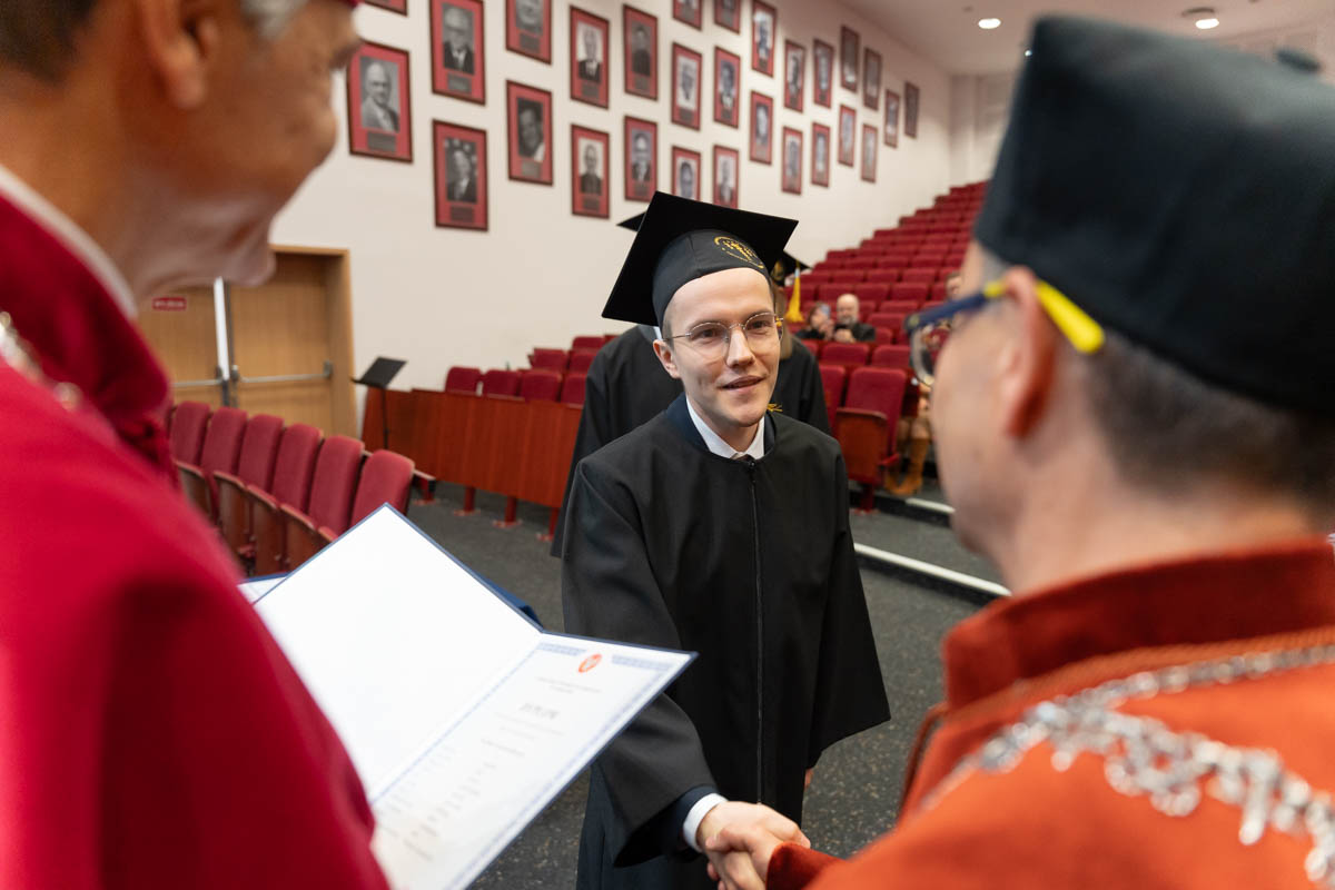 A person in a gown and mortarboard is shaking hands with another person dressed in formal attire during a ceremony in a hall with red seats. On the left side of the frame, another person is holding an open diploma. In the background, framed portraits hang on the wall.
