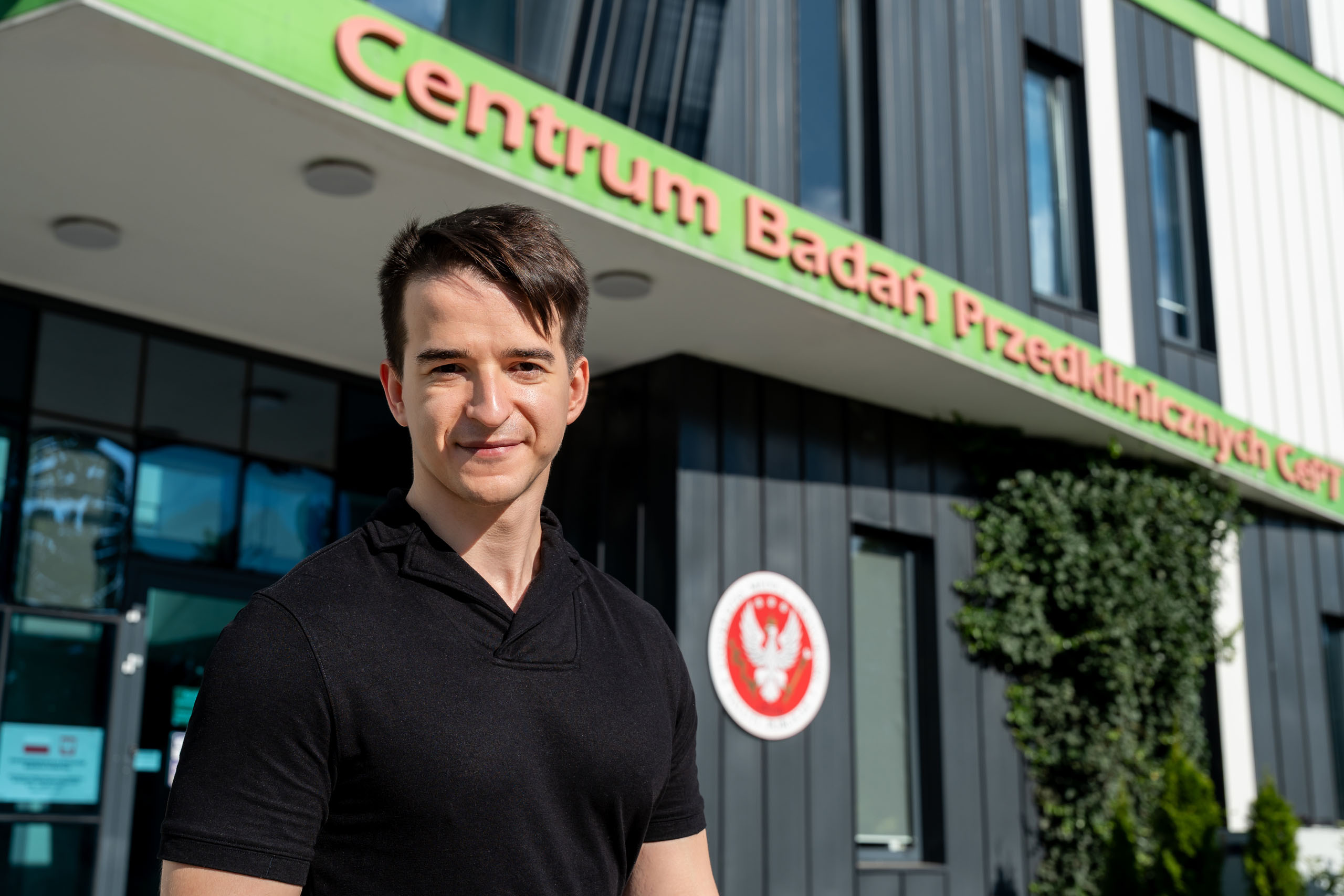 A person stands in front of a modern building with a green sign reading “Centrum Badań Przedklinicznych.” The person is wearing a black short-sleeved shirt and is positioned in the foreground, while the building façade, entrance, windows, and a red-and-white emblem are visible in the background.