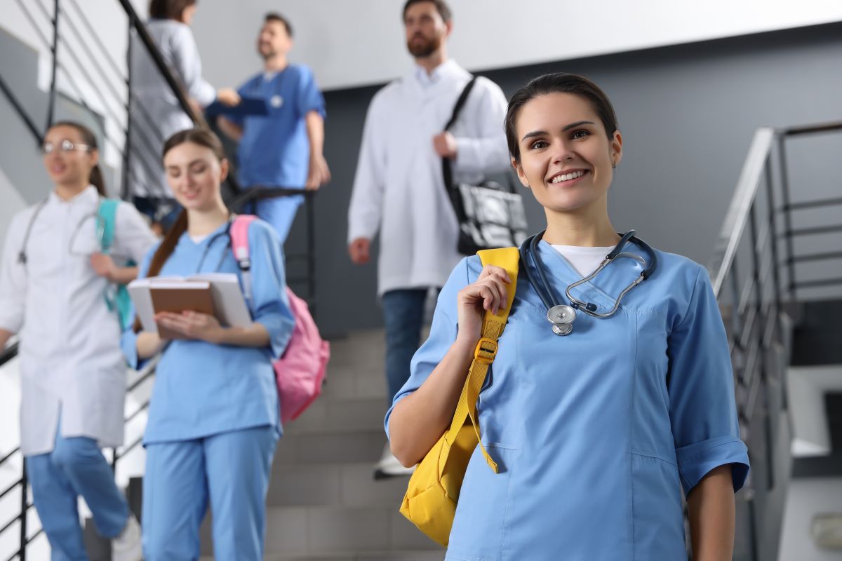 Na pierwszym planie osoba w niebieskim stroju medycznym ze stetoskopem na szyi, trzymająca na ramieniu żółtą torbę. W tle na klatce schodowej znajduje się grupa osób w strojach medycznych i roboczych, w tym osoby w niebieskich i białych uniformach. Jedna z osób trzyma notatnik lub książkę, inne mają torby lub plecaki. Wszyscy przemieszczają się wewnątrz nowoczesnego budynku o jasnych, szarych ścianach.