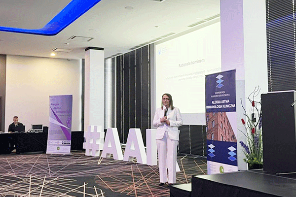 Kobieta stoi na środku sali konferencyjnej i przemawia. Po lewej duży napis „#AAI”. W tle ekran z prezentacją i roll‑upy z napisem „Alergia Astma Immunologia Kliniczna”. 