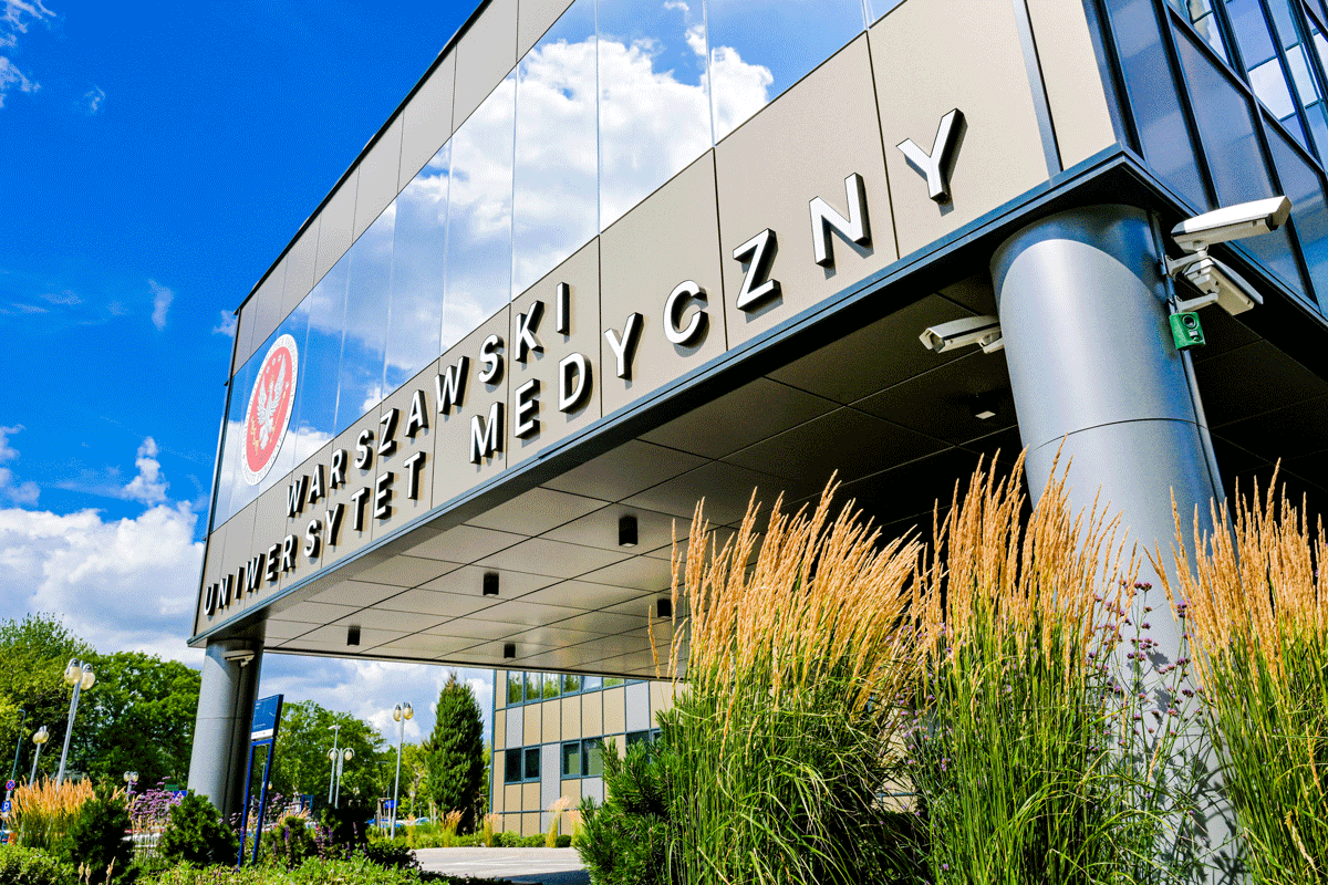 Budynek z przeszkloną fasadą. Na fasadzie litery tworzące napis WARSZAWSKI UNIWERSYTET MEDYCZNY oraz czerwono-białe godło WUM o okrągłym kształcie. Wokół budynku roślinność. 