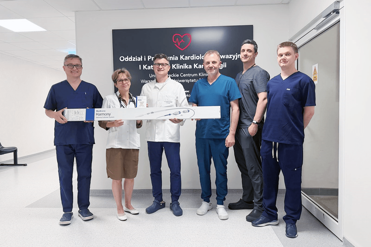 Sześć osób stoi w pomieszczeniu szpitalnym przy tablicy z napisem Oddział i Pracownia Kardiologii Inwazyjnej i Klinika Kardiologii. Osoby wspólnie trzymają długie pudełko z urządzeniem medycznym.