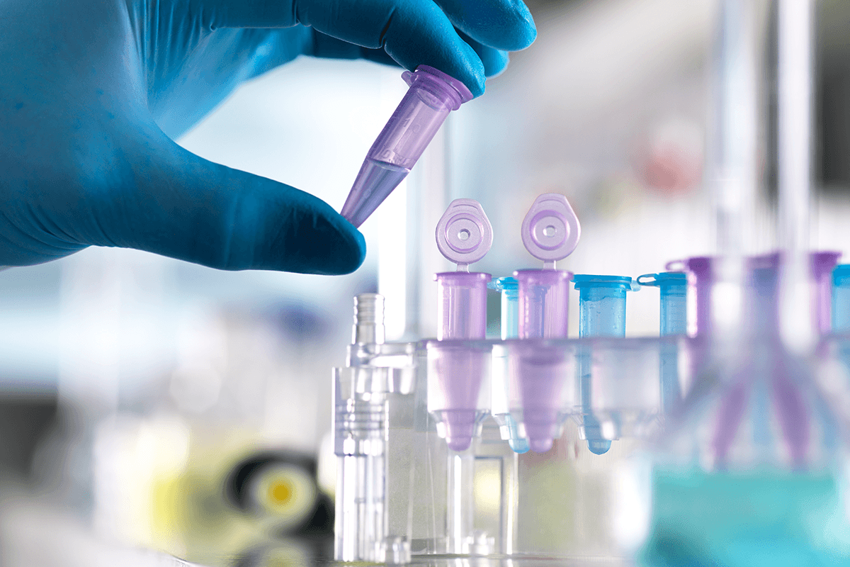 Dłoń w niebieskiej rękawiczce trzymająca tubkę laboratoryjną. Wokół dłoni widać inne przyrządy laboratoryjne.
