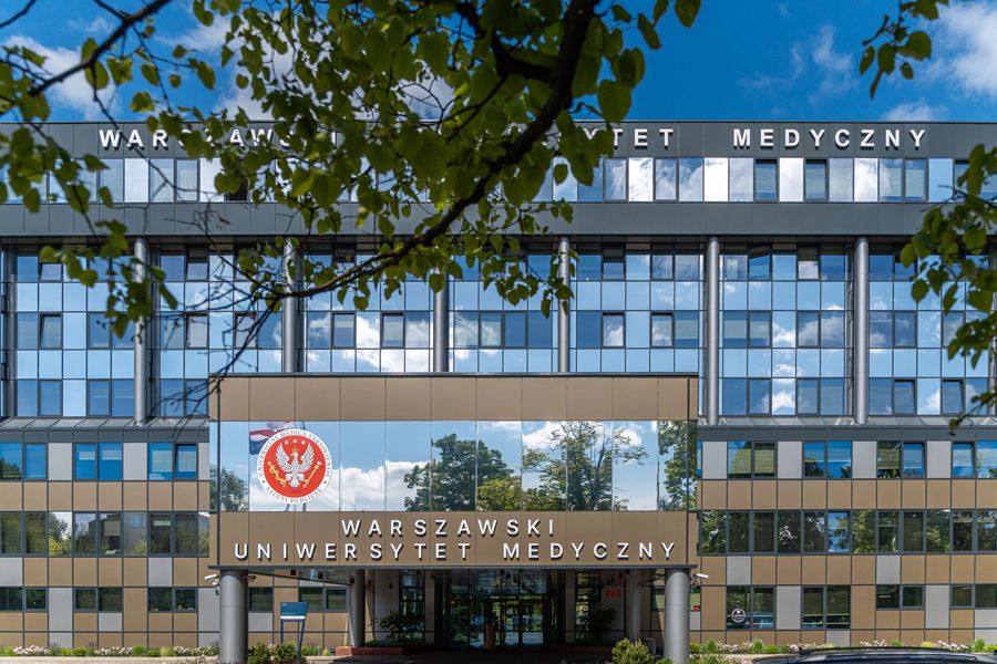 Building of the Medical University of Warsaw with a glass façade and main entrance. Above the entrance, the inscription “Medical University of Warsaw” and the university emblem are visible. On the upper part of the façade, another partially obscured inscription shows only a fragment: “… Medical University.” Trees stand in front of the building.