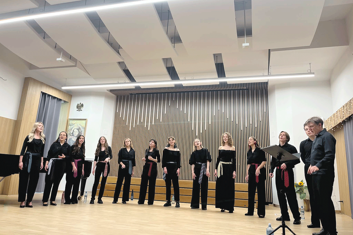 Grupa 12 osób ubranych na czarno stoi na scenie. Są w trakcie wykonywania utworu muzycznego.