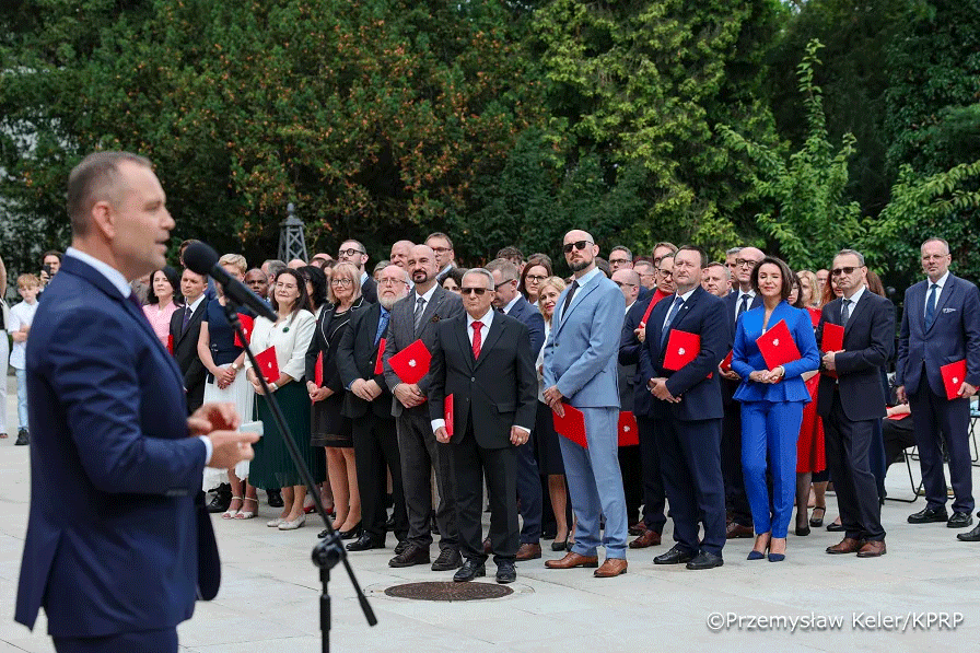 A group of people during a nomination ceremony. In the foreground on the left, the President is standing behind a microphone. In front of him are professors holding red folders in their hands.