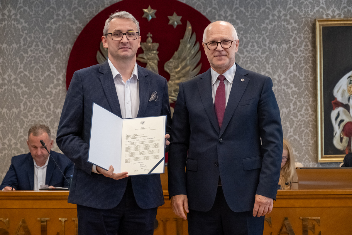 Dwie osoby stoją przed stołem prezydialnym. Jedna z nich trzyma otwartą teczkę z dokumentem skierowanym w stronę aparatu. W tle widoczne są osoby uczestniczące w posiedzeniu oraz godło na ścianie.
