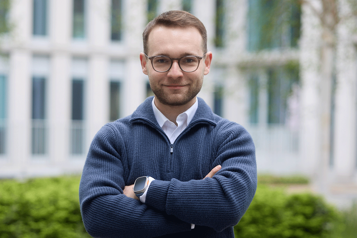 Młody mężczyzna pozuje do zdjęcia. W tle widać jasny budynek i zieleń.