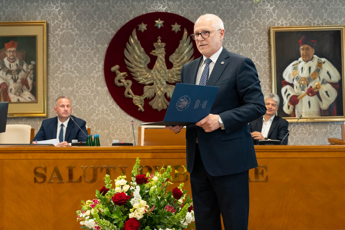 Jedna osoba stoi przed stołem prezydialnym i trzyma otwartą teczkę z dokumentem. W tle siedzą dwie osoby. Za uczestnikami widoczny jest godło oraz portrety na ścianie sali obrad.