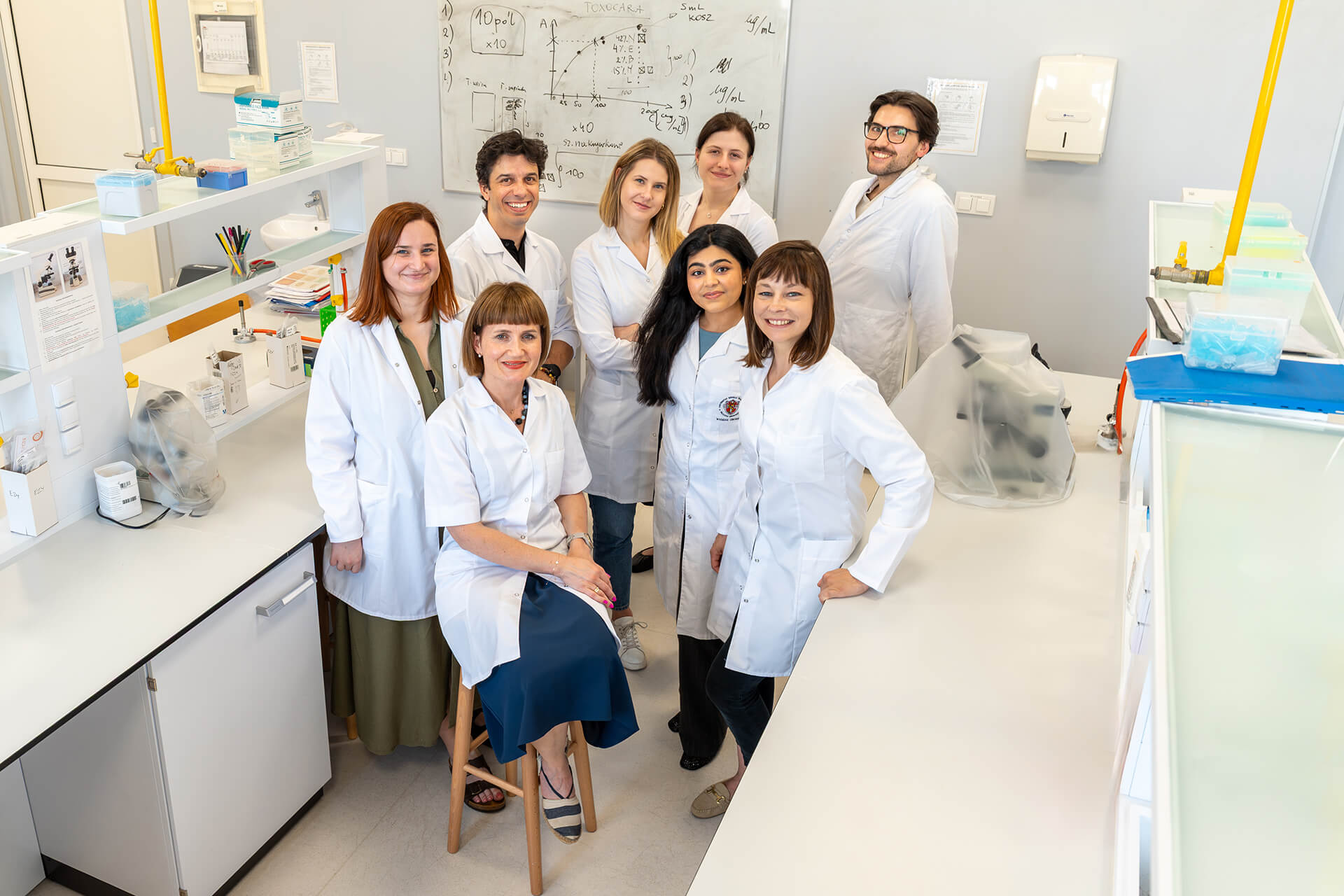 Grupa osób ubranych w fartuchy medyczne pozuje do wspólnego zdjęcia w pomieszczeniu laboratoryjnym.