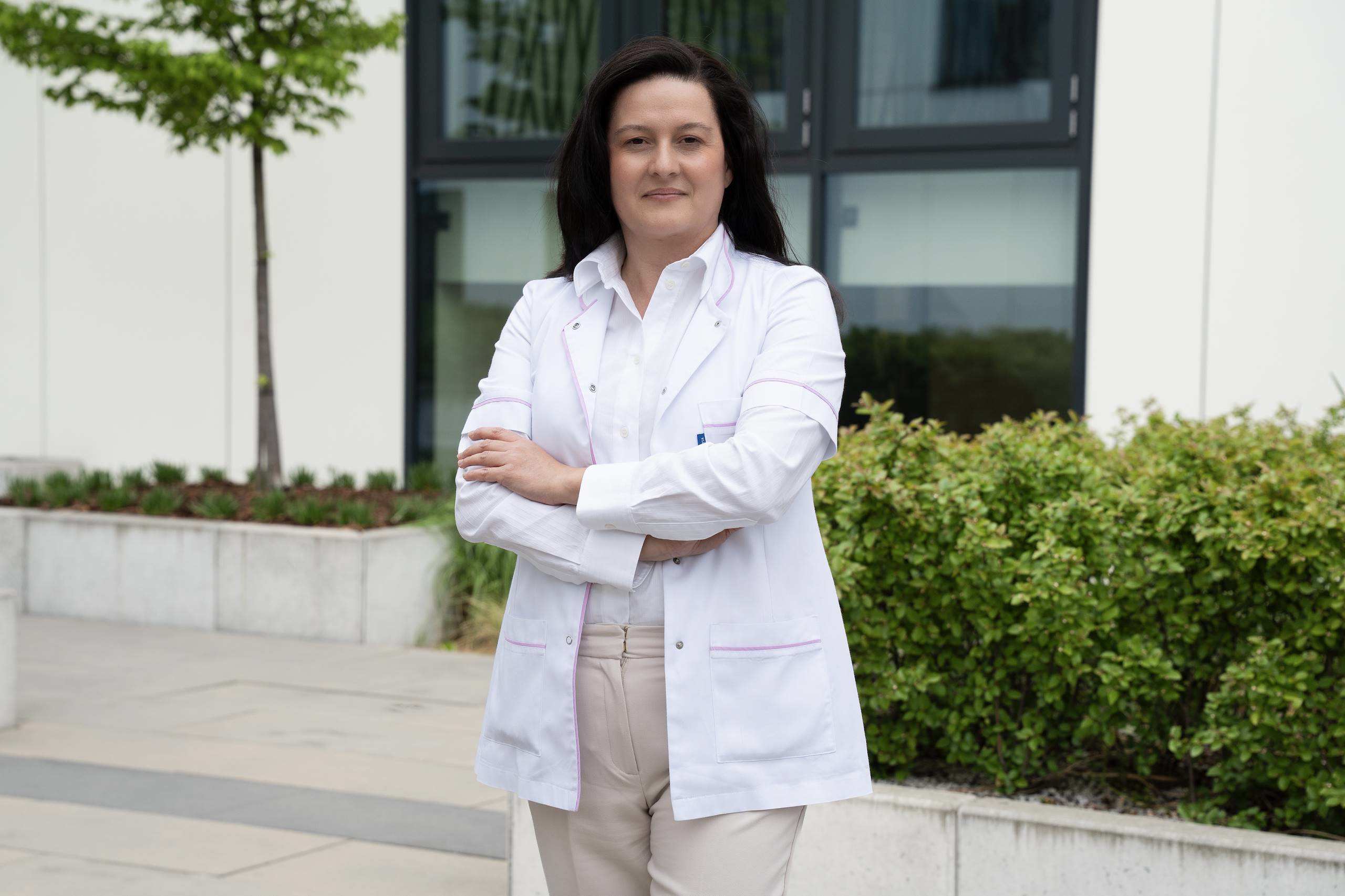 A woman wearing a white medical scrub poses for a photo against a backdrop of greenery and a building with a light-colored facade.