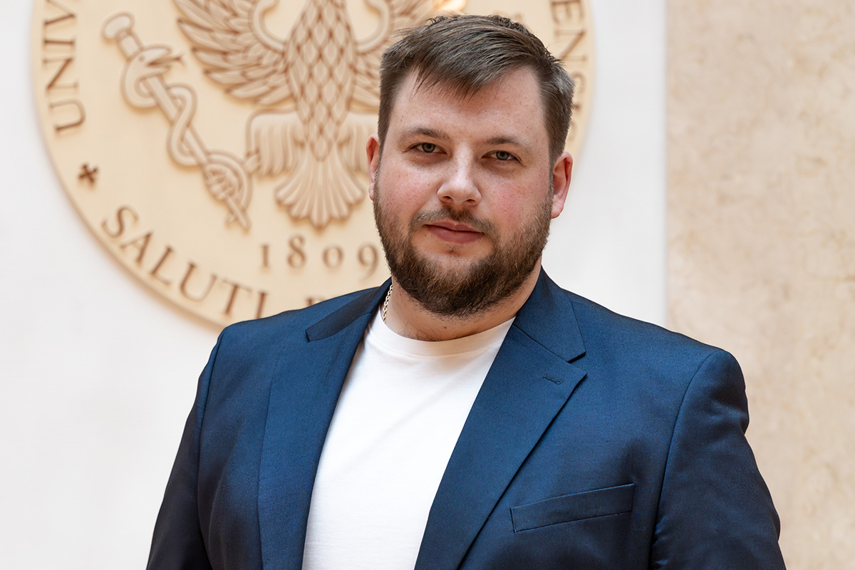 Młody mężczyzna, Adam Bujanowicz, pozuje do zdjęcia na tle godła WUM.