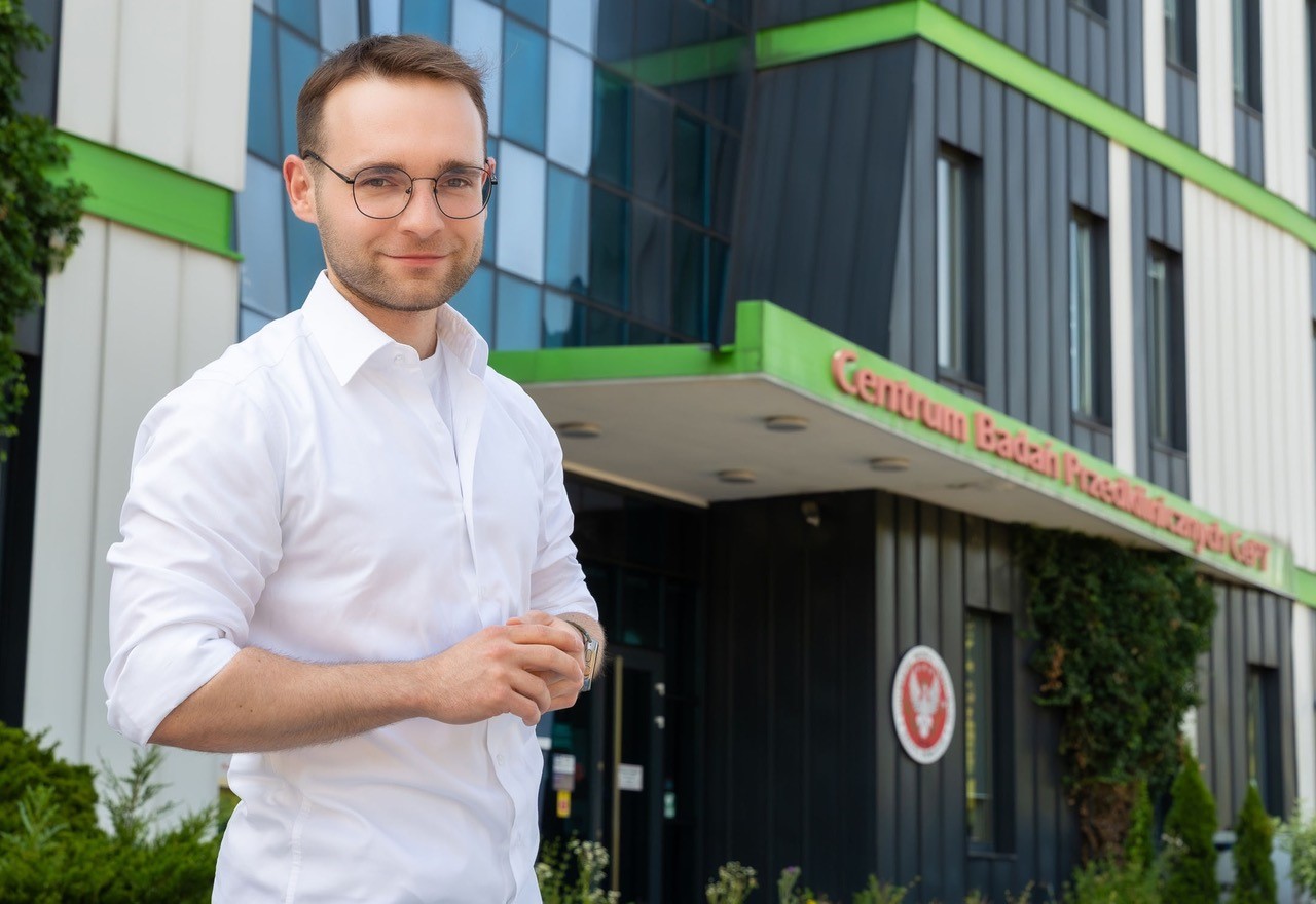 A young man, Karol Momot, poses for a photo in front of a building bearing the sign “Preclinical Research Center”.