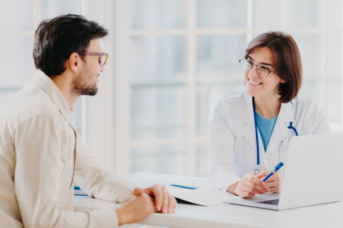 Healthcare professional in a white coat consulting with a patient across a desk in a bright medical office, with a laptop and documents visible.