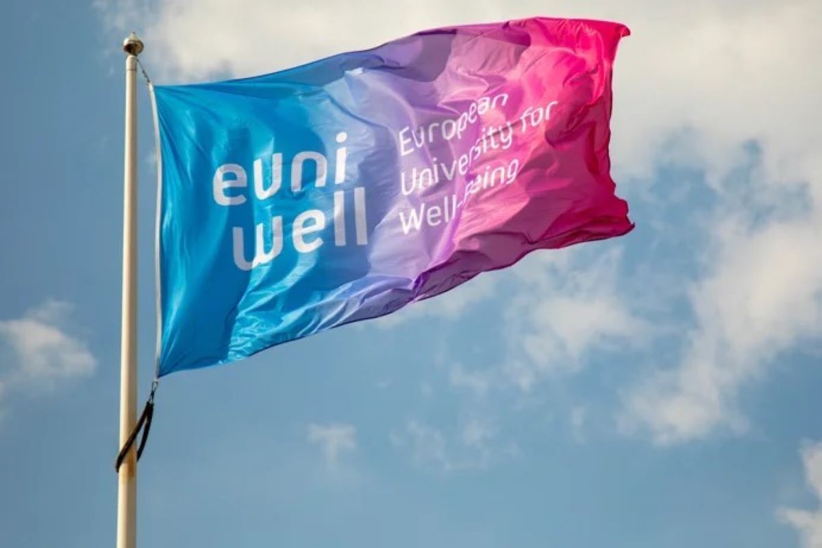 EUniWell flag in blue, purple, and pink colors waving on a flagpole against a partly cloudy sky. The flag displays the text ‘European University for Well-Being