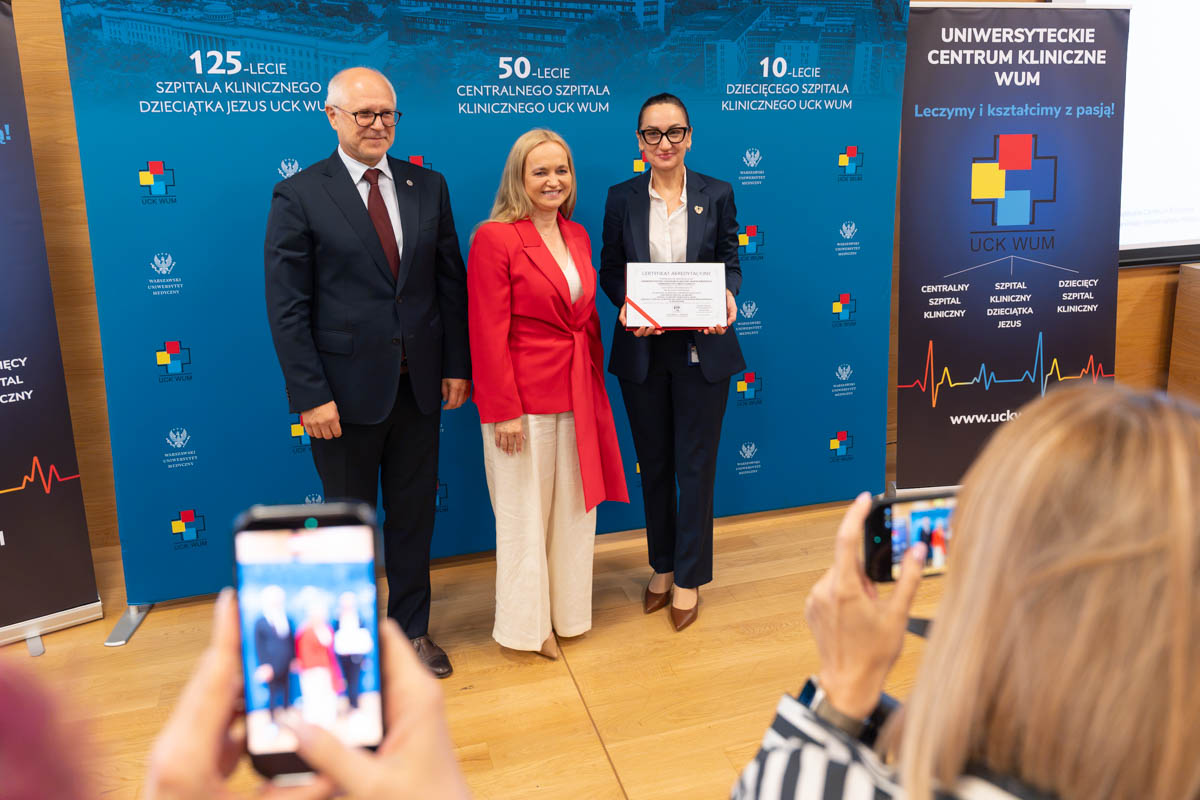 Trzy osoby stoją przed ścianką Uniwersyteckiego Centrum Klinicznego WUM. Jedna z osób trzyma certyfikat, a uczestnicy wydarzenia fotografują moment wręczenia dokumentu.