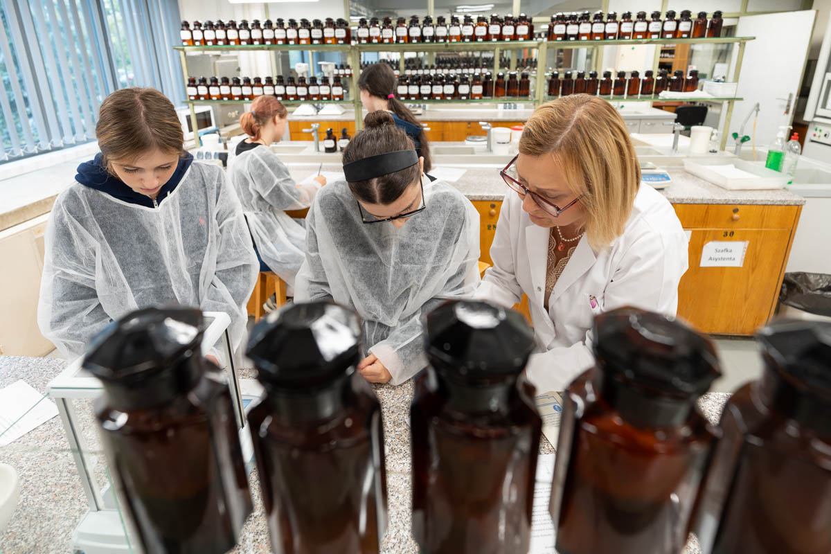 Osoby uczestniczące w zajęciach laboratoryjnych pracują przy stole laboratoryjnym pod nadzorem prowadzącej. Na pierwszym planie widoczne są szklane butelki z odczynnikami.