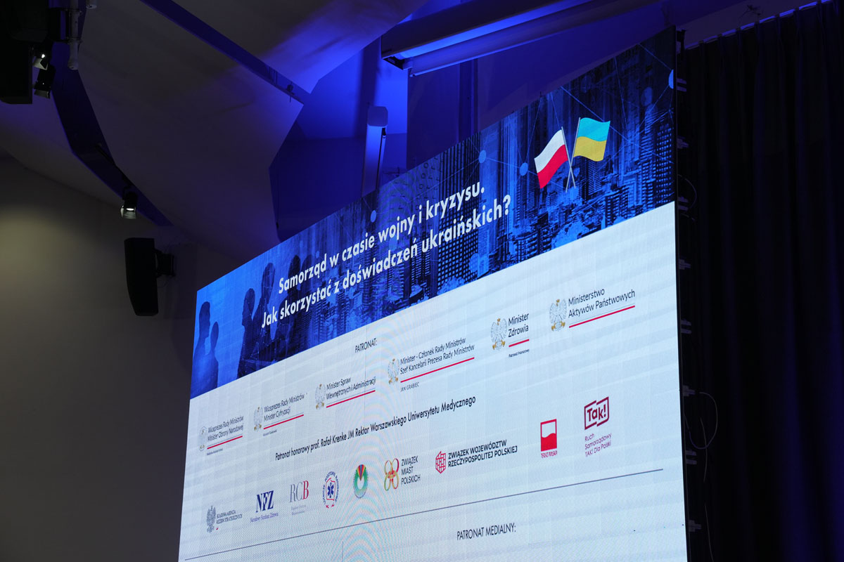 Ekran projekcyjny prezentuje planszę konferencji „Samorząd w czasie wojny i kryzysu. Jak skorzystać z doświadczeń ukraińskich?”. W górnej części widoczne są flagi Polski i Ukrainy. Na planszy znajdują się logotypy patronów, partnerów i patronów medialnych wydarzenia. Zdjęcie wykonano w sali konferencyjnej.