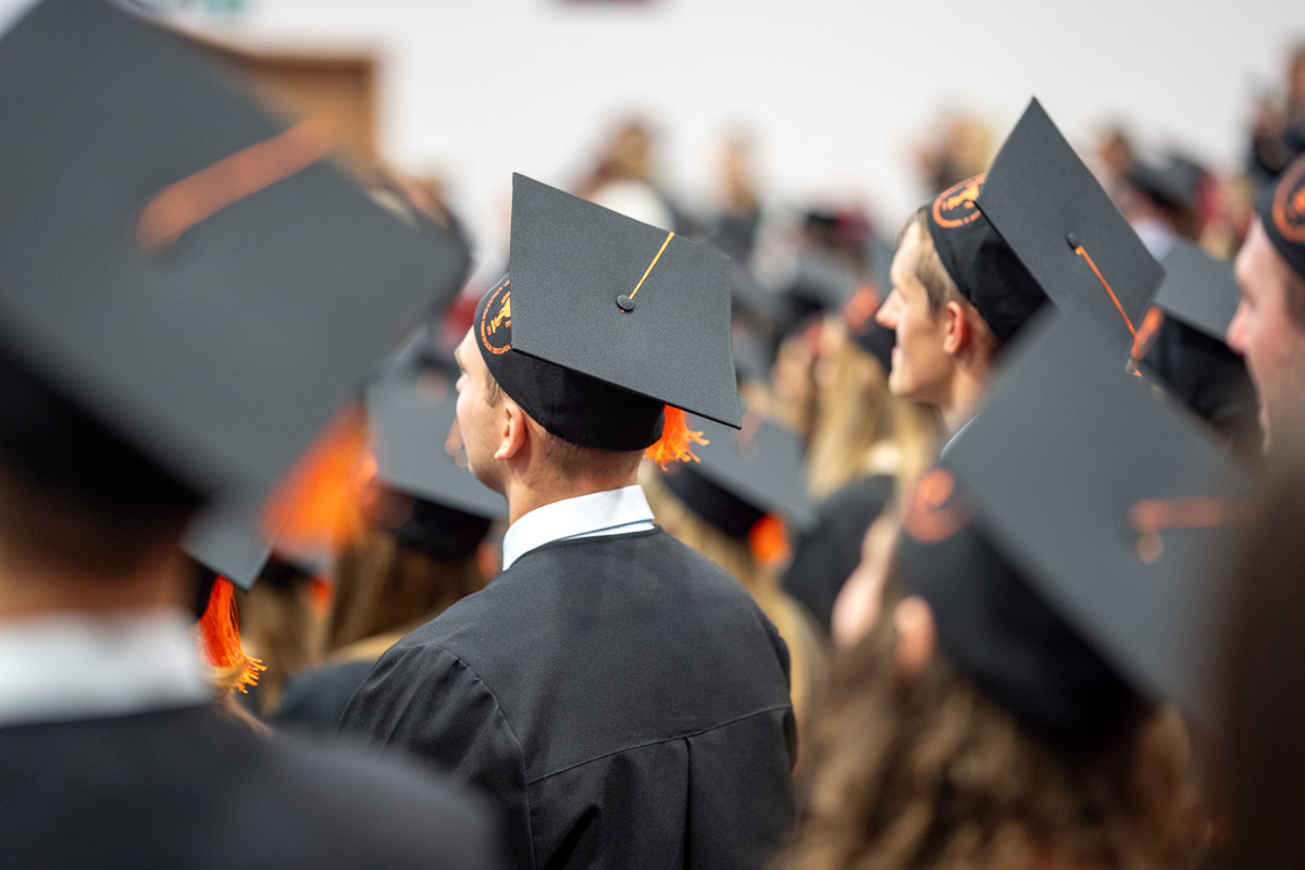 Grupa osób w togach i biretach uczestniczy w uroczystości ukończenia studiów. W centrum kadru widoczna jest osoba stojąca tyłem, a wokół znajdują się pozostali uczestnicy ceremonii. W tle widać zgromadzoną publiczność.