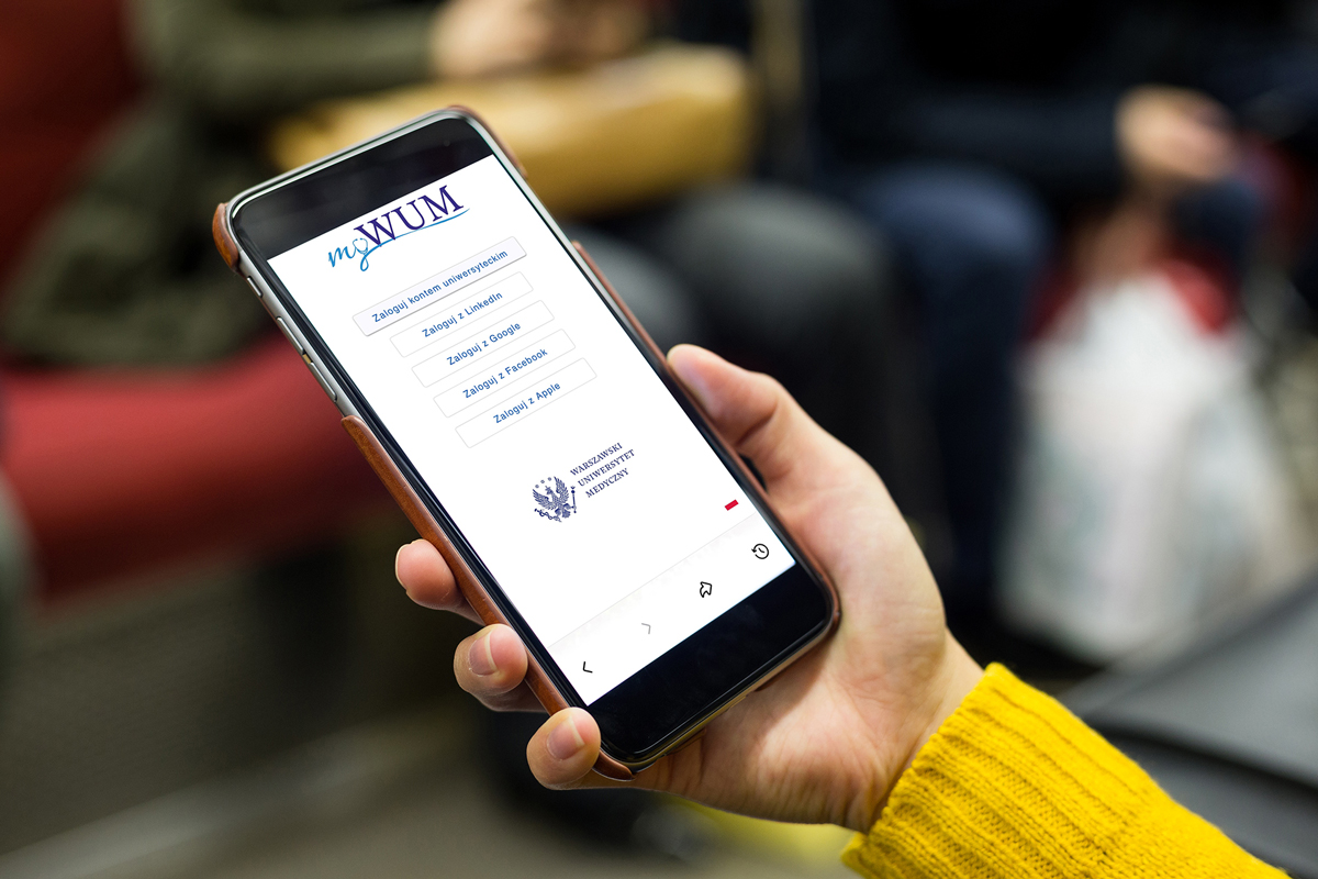 Osoba trzyma smartfon z otwartą stroną logowania do systemu myWUM. Na ekranie widoczne są przyciski logowania kontem uniwersyteckim oraz przez LinkedIn, Google, Facebook i Apple. W dolnej części ekranu znajduje się logo Warszawskiego Uniwersytetu Medycznego. Tło jest nieostre.