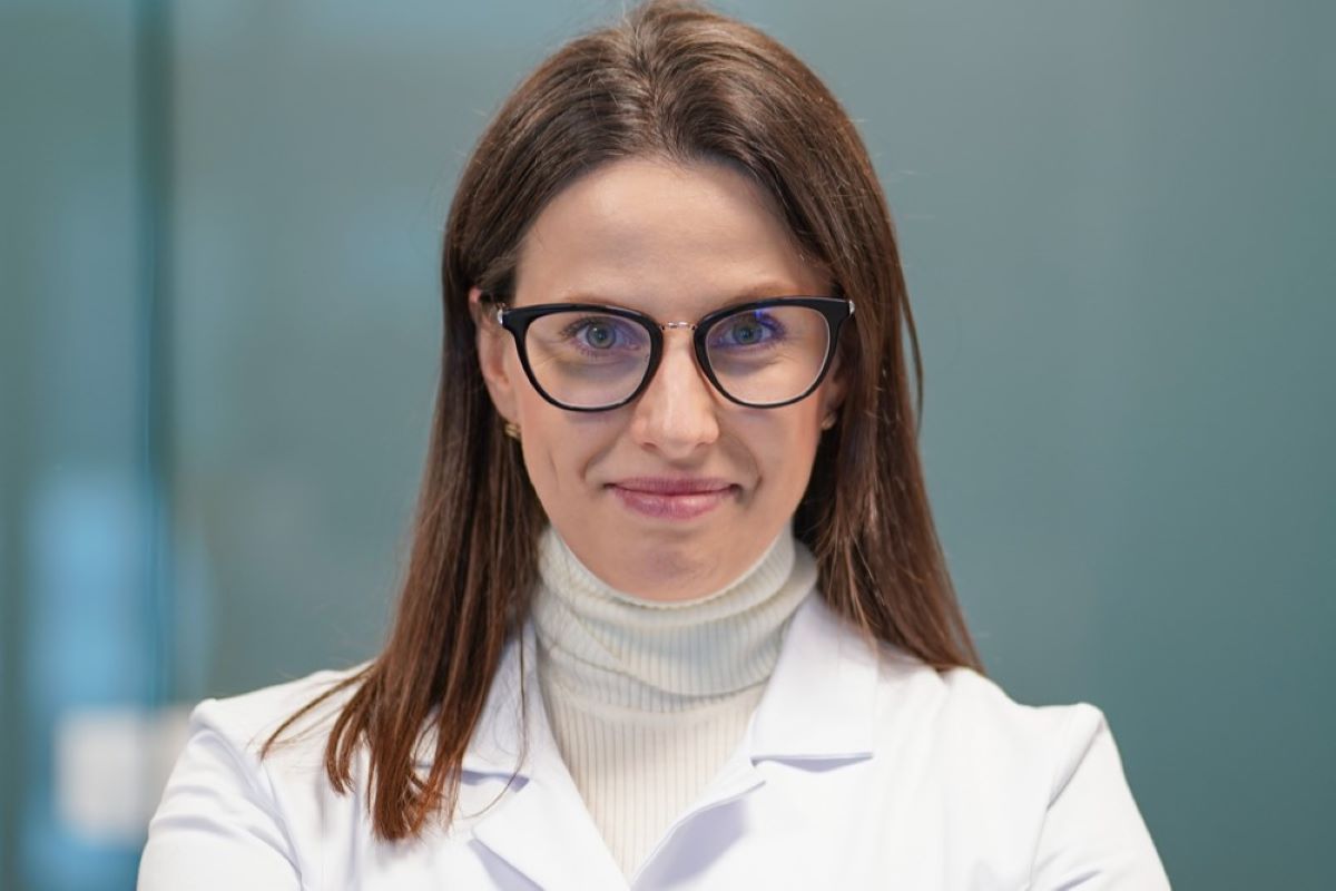 Portret osoby w białym fartuchu i ciemnych okularach, z długimi włosami, na tle rozmytego wnętrza. Osoba patrzy w obiektyw.