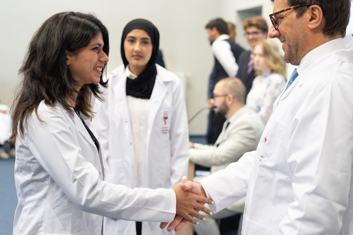 Młoda kobieta i mężczyzna w białych fartuchach podają sobie ręce podczas ceremonii White Coat Ceremony. W tle stoi kolejna osoba uczestnicząca w wydarzeniu, a przy stanowisku obsługi widoczne są dwie osoby.