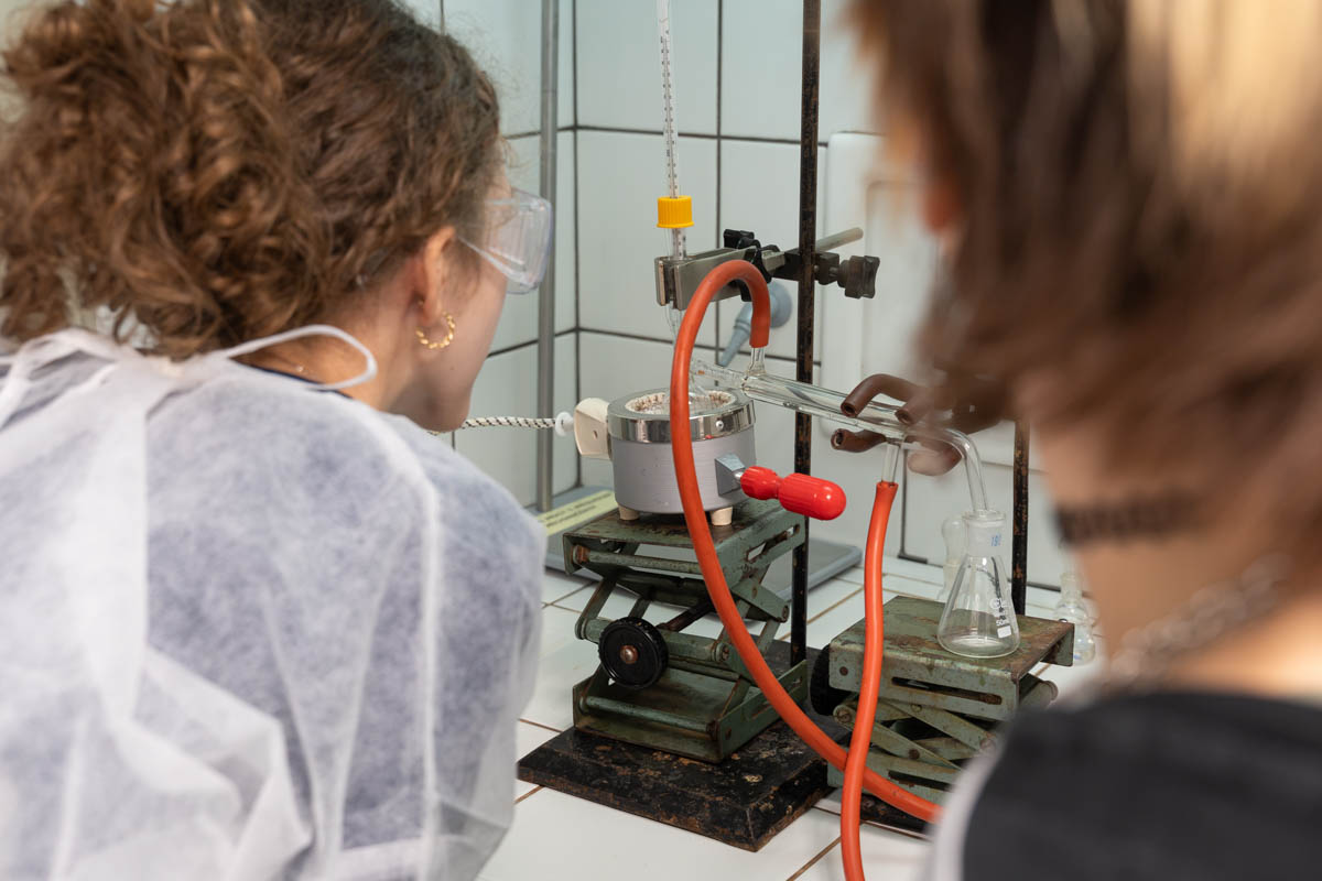 Dwie osoby obserwują doświadczenie laboratoryjne. Na stanowisku znajduje się szklana aparatura połączona przewodami oraz kolba ustawiona pod chłodnicą. Jedna osoba nosi okulary ochronne i odzież laboratoryjną.