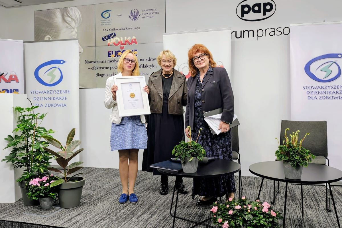 Trzy osoby stoją na scenie podczas konferencji „Polka w Europie”. Jedna osoba trzyma oprawiony dyplom. W tle widoczne są banery Stowarzyszenia Dziennikarze dla Zdrowia, Warszawskiego Uniwersytetu Medycznego oraz PAP. Przed uczestnikami stoją stoliki i donice z roślinami.