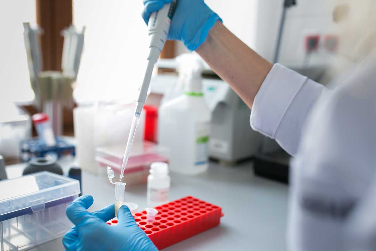 Osoba wykonuje czynności laboratoryjne przy użyciu pipety automatycznej. W dłoni trzyma probówkę, do której przenosi próbkę. Na blacie znajdują się stojak na probówki oraz wyposażenie laboratoryjne.