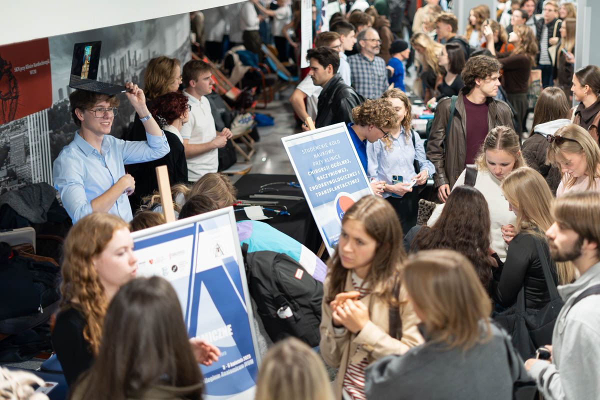 A crowd of participants at the Student Research Groups Fair gathers around stands showcasing the groups’ activities. One person is holding an open laptop above their head, while others talk and browse the materials on display.