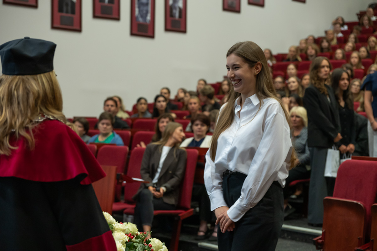 A person in a white shirt stands opposite a person wearing a red academic gown and a black mortarboard. In the background, ceremony attendees are seated in rows of red chairs.