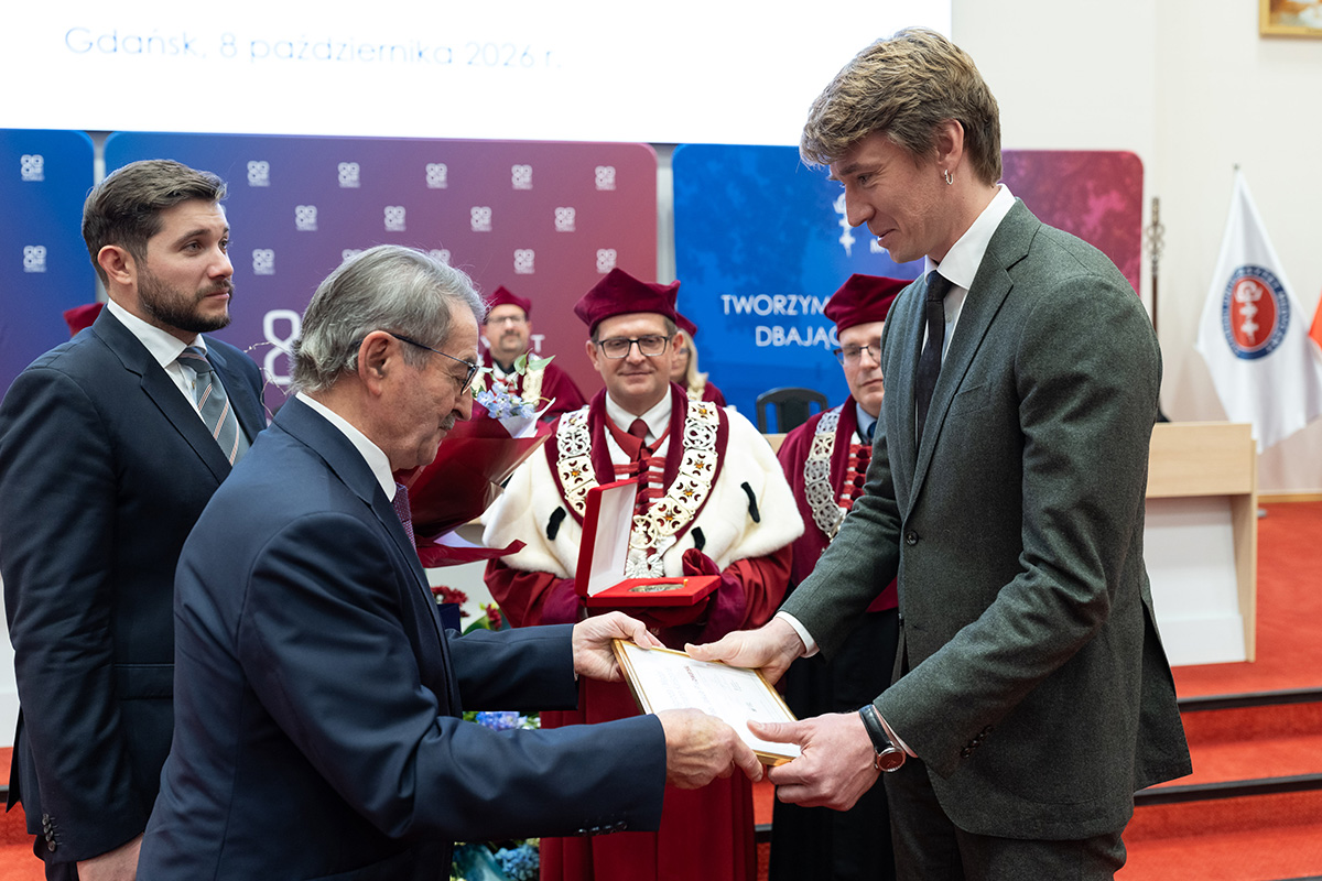 Mężczyzna, profesor Piwowarski (po prawej) odbiera medal i dyplom od innego mężczyzny. Obok stoją trzej mężczyźni, dwaj w strojach rektorskich. W tle widać kolorową ściankę z logiem i nazwą Gdańskiego Uniwersytetu Medycznego.