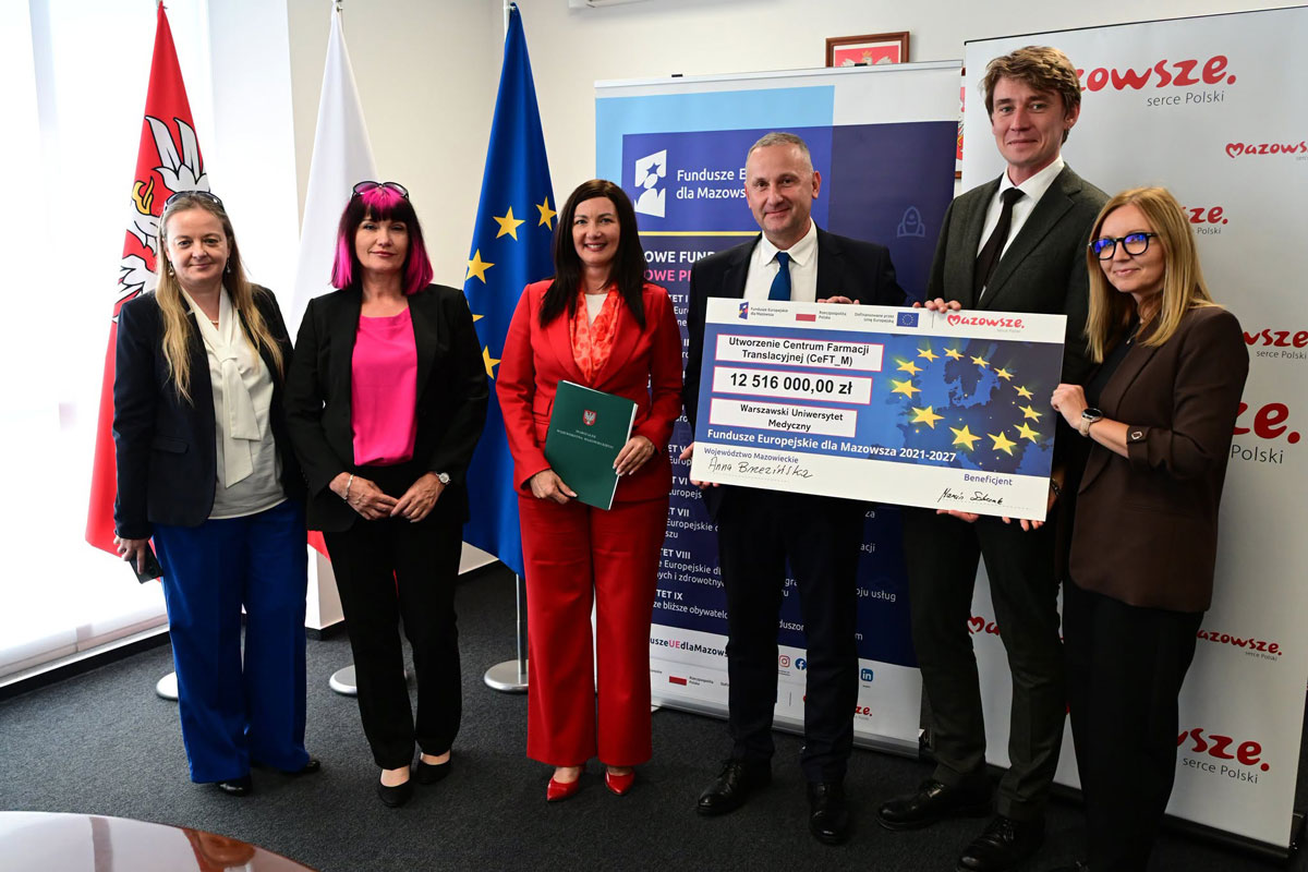 Sześć osób stoi podczas oficjalnego wydarzenia. Dwie osoby trzymają dużą planszę z informacją o dofinansowaniu projektu. W tle widoczne są flagi Polski i Unii Europejskiej oraz materiały promujące Fundusze Europejskie dla Mazowsza.