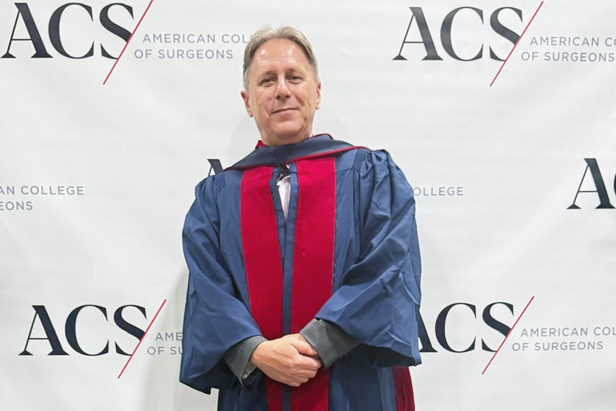 Mężczyzna profesor Myrcha stoi przed ścianką z logo ACS (American College of Surgeons). Ma na sobie strój akademicki i patrzy w kierunku obiektywu. W tle wielokrotnie powtarza się nazwa organizacji.