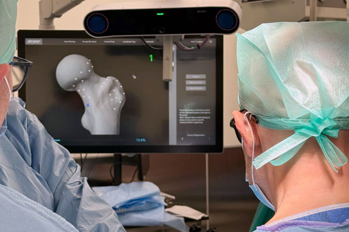 Dwóch członków zespołu operacyjnego wykonuje zabieg z wykorzystaniem systemu robotycznego. Na monitorze widoczny jest trójwymiarowy model stawu biodrowego używany do planowania i nawigacji podczas operacji.