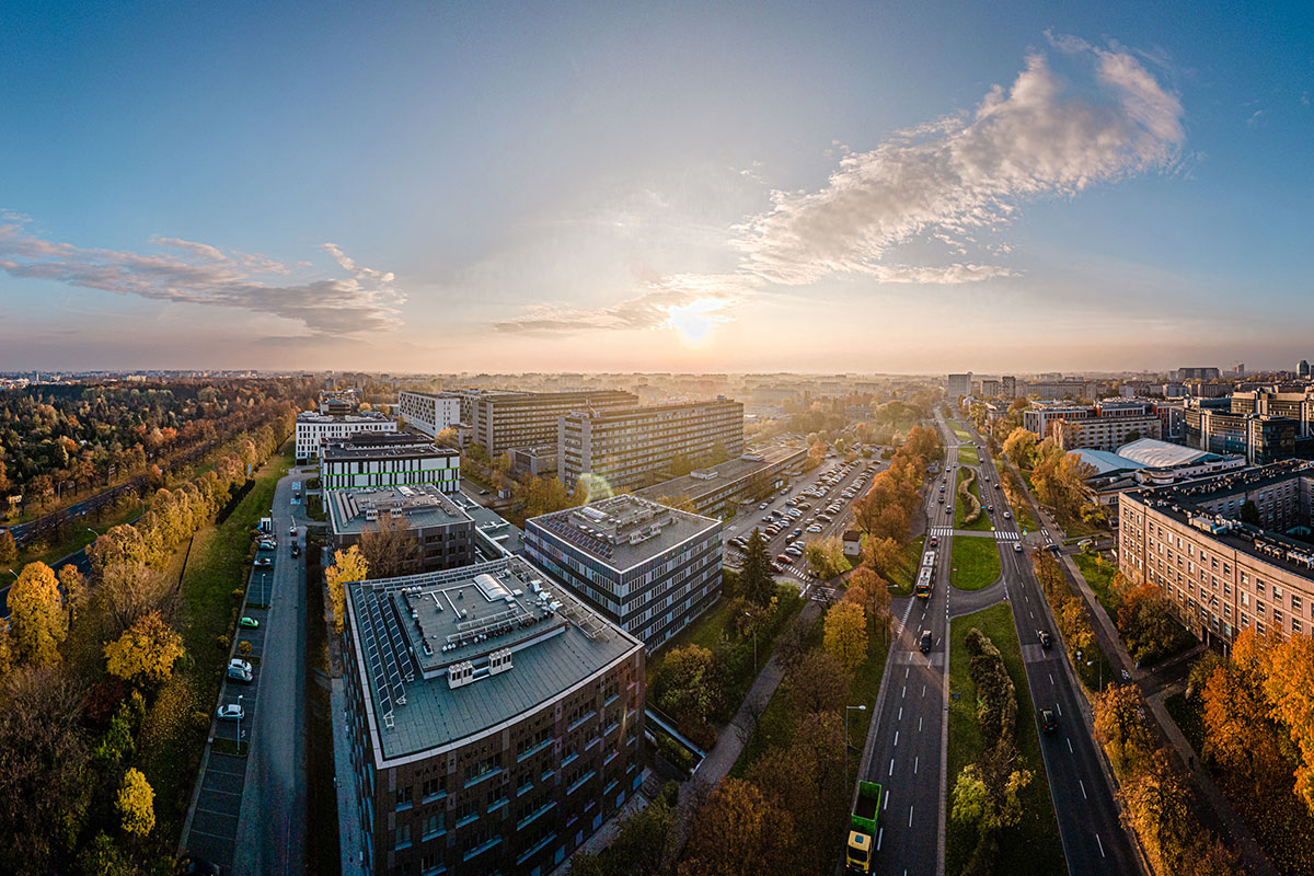 Panorama kampusu z lotu ptaka. W centrum znajdują się nowoczesne budynki biurowo-dydaktyczne, a po prawej biegnie szeroka wielopasmowa ulica z torowiskiem tramwajowym i samochodami. Po lewej stronie widoczny jest pas zieleni z drzewami. W tle rozciąga się zabudowa miasta. Nad horyzontem znajduje się nisko położone słońce i częściowo zachmurzone niebo.