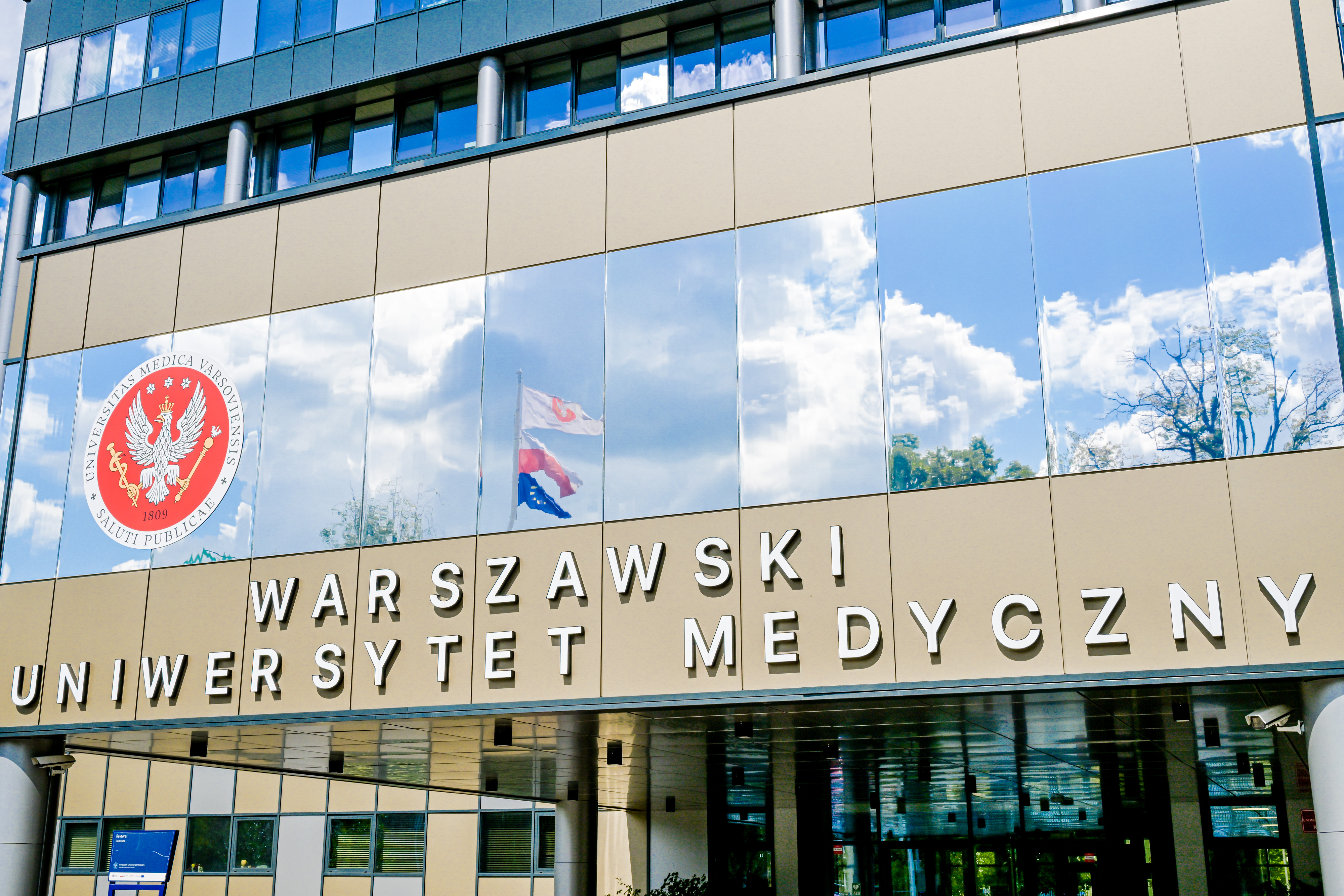 Fasada budynku Warszawskiego Uniwersytetu Medycznego z nazwą uczelni i godłem na elewacji; w szklanych oknach odbijają się niebo, chmury i drzewa.