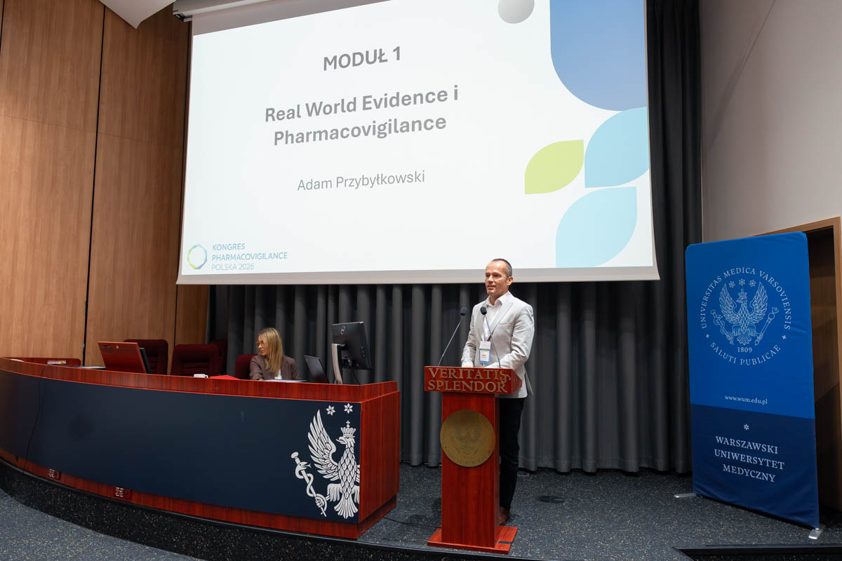 Jedna osoba przemawia przy mównicy z dwoma mikrofonami podczas Kongresu Pharmacovigilance Polska 2026. Obok stoi baner Warszawskiego Uniwersytetu Medycznego.
