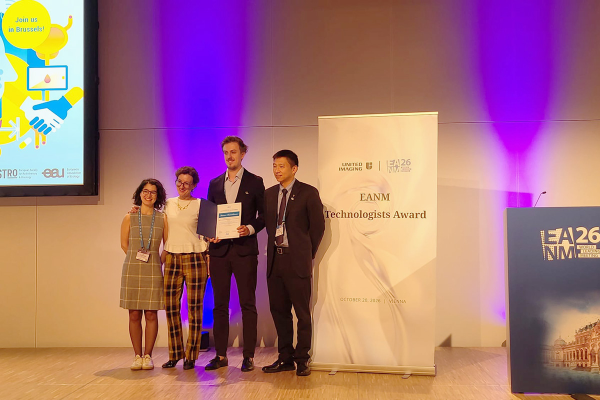 Cztery osoby stoją na scenie podczas wręczenia nagrody EANM Technologists Award. Jedna osoba trzyma otwarty dyplom. Obok znajduje się roll-up z nazwą nagrody i logotypami wydarzenia EANM 2026. W tle widoczna jest podświetlona ściana oraz fragment ekranu prezentacyjnego.