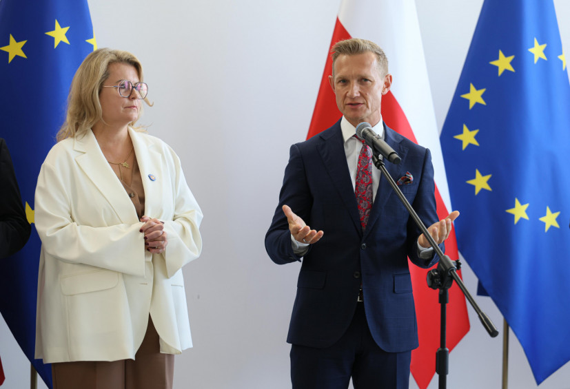 Dwie osoby stoją przed mikrofonem podczas oficjalnego wydarzenia. Jedna osoba przemawia, druga stoi obok. W tle widoczne są flagi Polski i Unii Europejskiej.