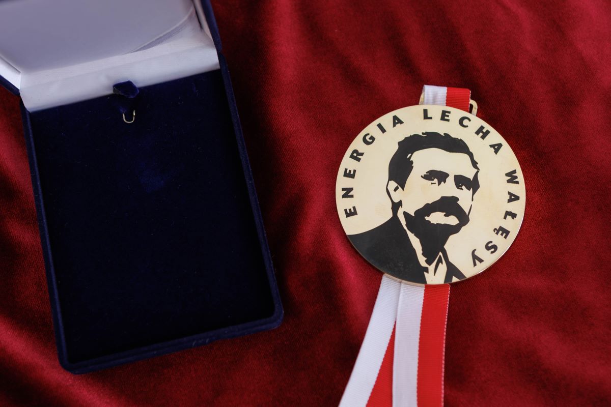Medal z wizerunkiem i napisem „Energia Lecha Wałęsy” leżący na czerwonym materiale obok otwartego granatowego etui wyłożonego ciemnym welurem. Medal jest zawieszony na biało-czerwonej wstążce.