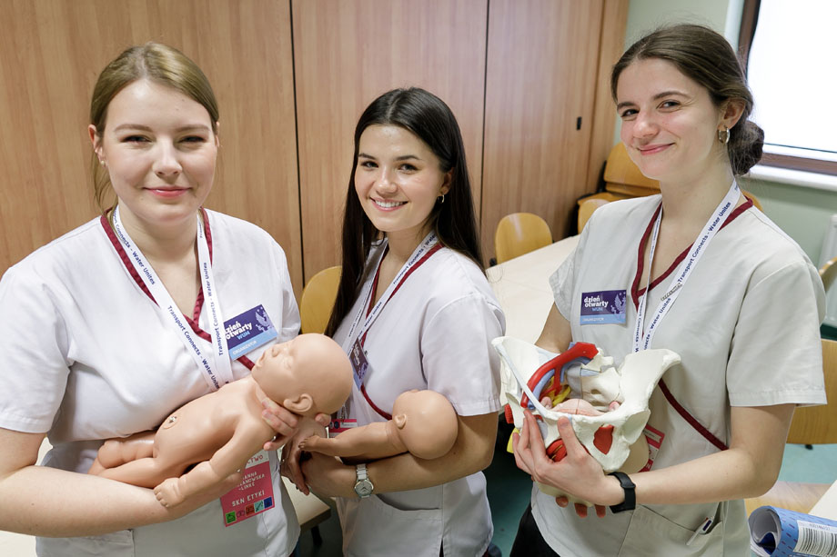 Trzy młode dziewczyny ubrane w fartuchy medyczne. Trzymają lalki noworodków. Uśmiechają się do aparatu.