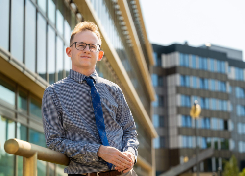 Dr hab. Piotr Alster, młody mężczyzna w okularach, ubrany w koszulę i krawat pozuje do zdjęcia na tle budynków z przeszklonymi oknami. Uśmiecha się.
