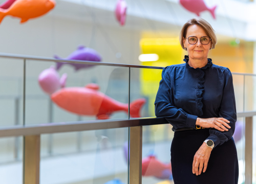 Kobieta stoi przy szklanej balustradzie w nowoczesnym wnętrzu, w tle zawieszone kolorowe dekoracje w kształcie ryb. Ubrana jest w granatową bluzkę z falbaną i ciemną spódnicę, na nadgarstku ma zegarek typu smartwatch.