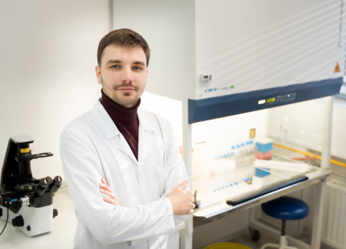 Młody mężczyzna ubrany w biały fartuch lekarski stoi w pomieszczeniu laboratoryjnym i pozuje do zdjęcia. W tle widać sprzęty medyczne.