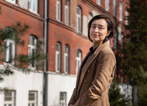 Osoba stojąca na zewnątrz, ubrana w elegancki płaszcz, na tle ceglanego budynku z dużymi oknami i otaczającej zieleni.