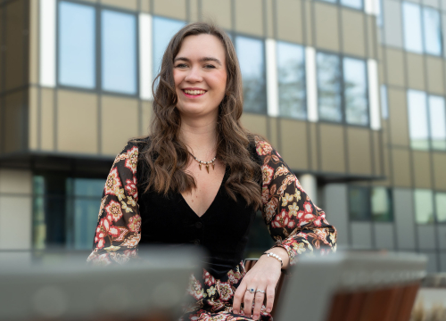 Młoda kobieta siedzi na ławce przed nowoczesnym budynkiem z dużymi przeszkleniami. Patrzy w stronę aparatu, uśmiecha się. Jedną rękę opiera rękę na oparciu ławki.