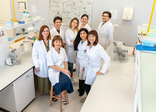Osiem osób ubranych w białe fartuchy pozuje do wspólnego zdjęcia w przestrzeni laboratoryjnej.